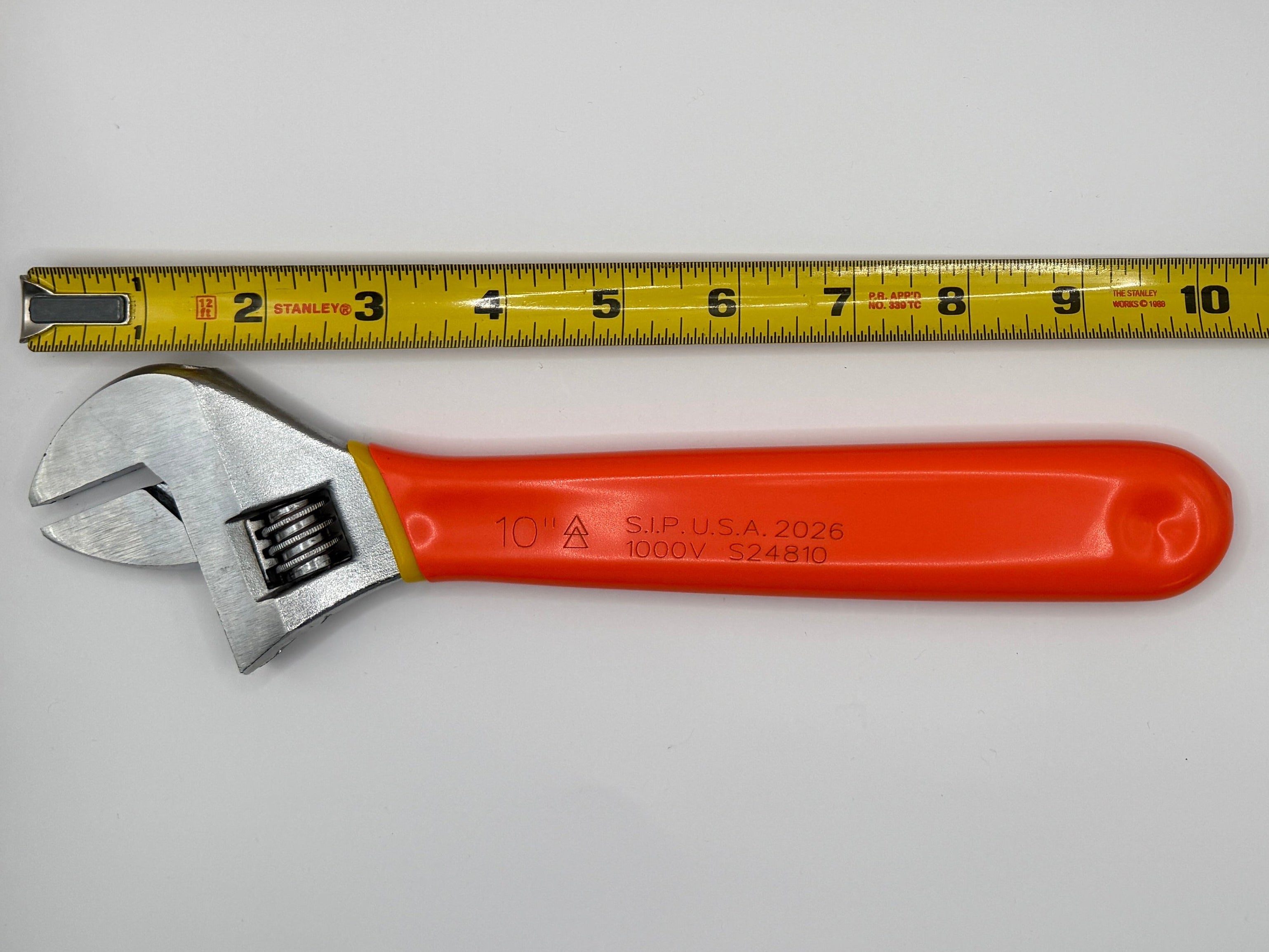 A yellow tape measure standing vertically next to an adjustable wrench with a bright orange handle, photographed inside a white box with a textured gray floor and ceiling.