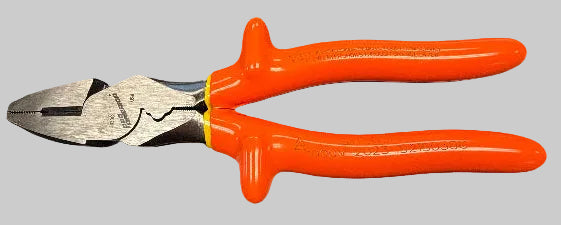 A pair of wire cutting pliers with bright orange handles and silver metal jaws, resting on a dark gray desk surface. Papers with handwritten notes and a calendar are visible in the background, and a person's lap is visible at the bottom of the frame.